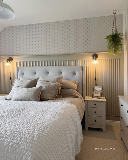 A styled bedroom interior featuring a bed dressed in light beige and gray bedding, a tufted headboard, ribbed half-height wall panelling, wall-mounted lamps, a hanging plant, and a bedside nightstand.