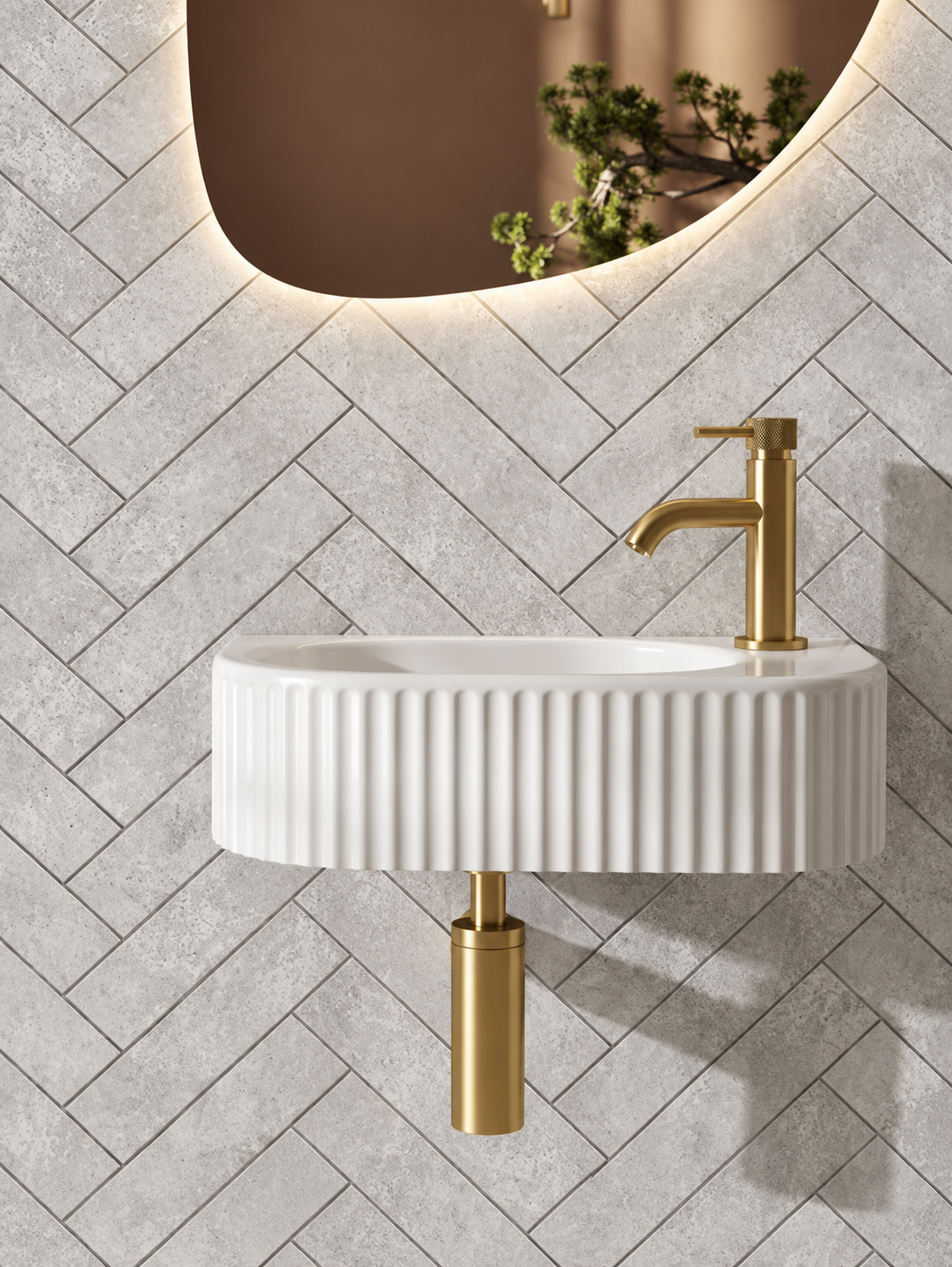 A modern wall-mounted bathroom sink with a ribbed white ceramic basin and a brushed gold faucet, installed against a gray herringbone tile wall under a curved mirror.