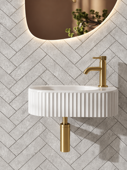 A modern wall-mounted bathroom sink with a ribbed white ceramic basin and a brushed gold faucet, installed against a gray herringbone tile wall under a curved mirror.