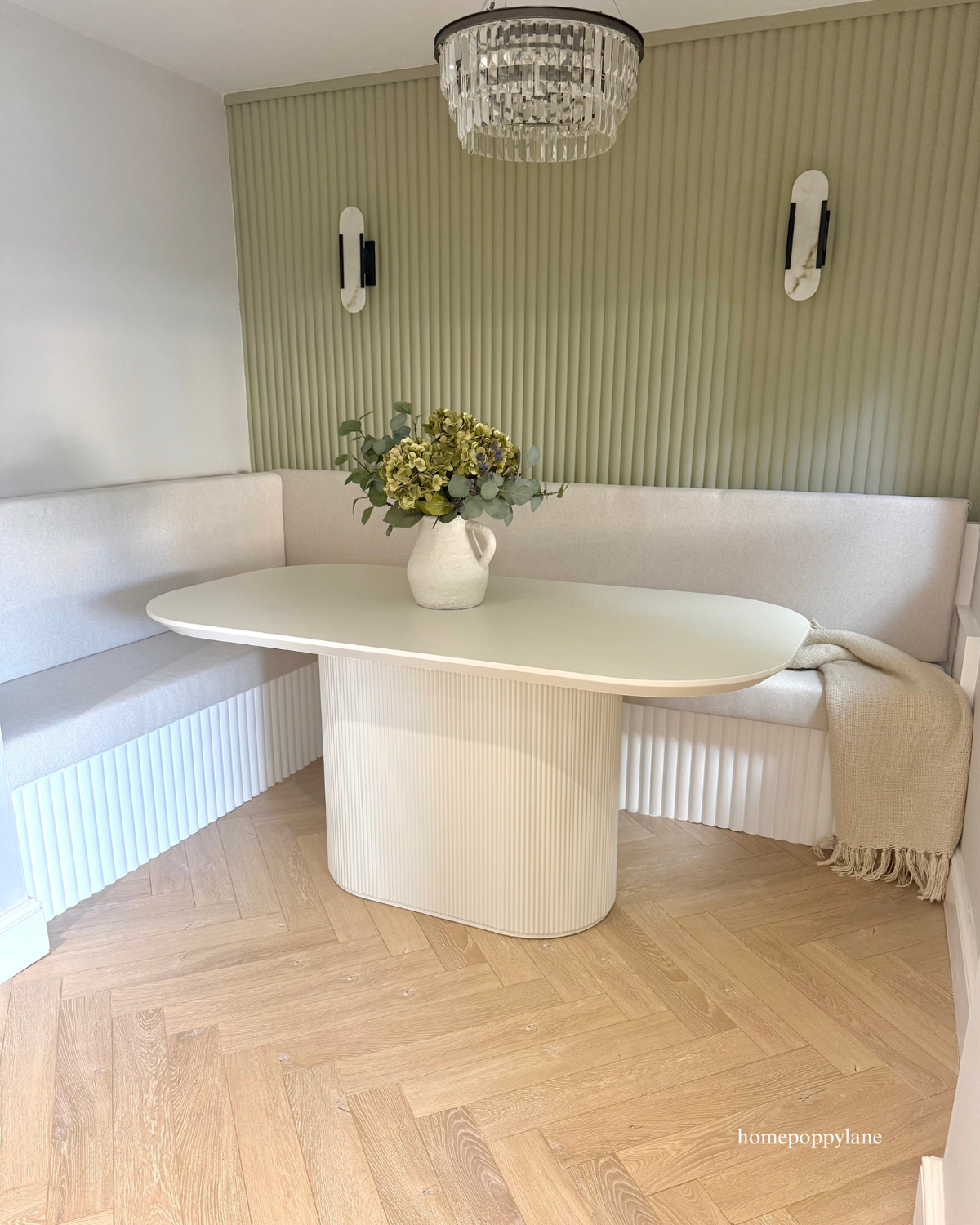 A modern white ribbed oval dining table in a bright corner booth with a light wood herringbone floor, a green ribbed accent wall, and a hanging crystal chandelier.