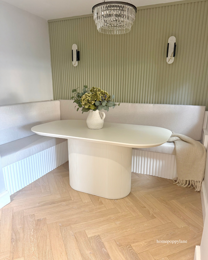 A modern white ribbed oval dining table in a bright corner booth with a light wood herringbone floor, a green ribbed accent wall, and a hanging crystal chandelier.