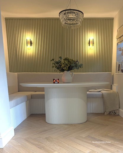 A modern dining nook featuring a white ribbed oval table, a matching white bench with a draped throw blanket, and a light wood slatted back wall illuminated by two wall sconces. A crystal chandelier hangs above, and green ribbed wall panels feature on the back wall