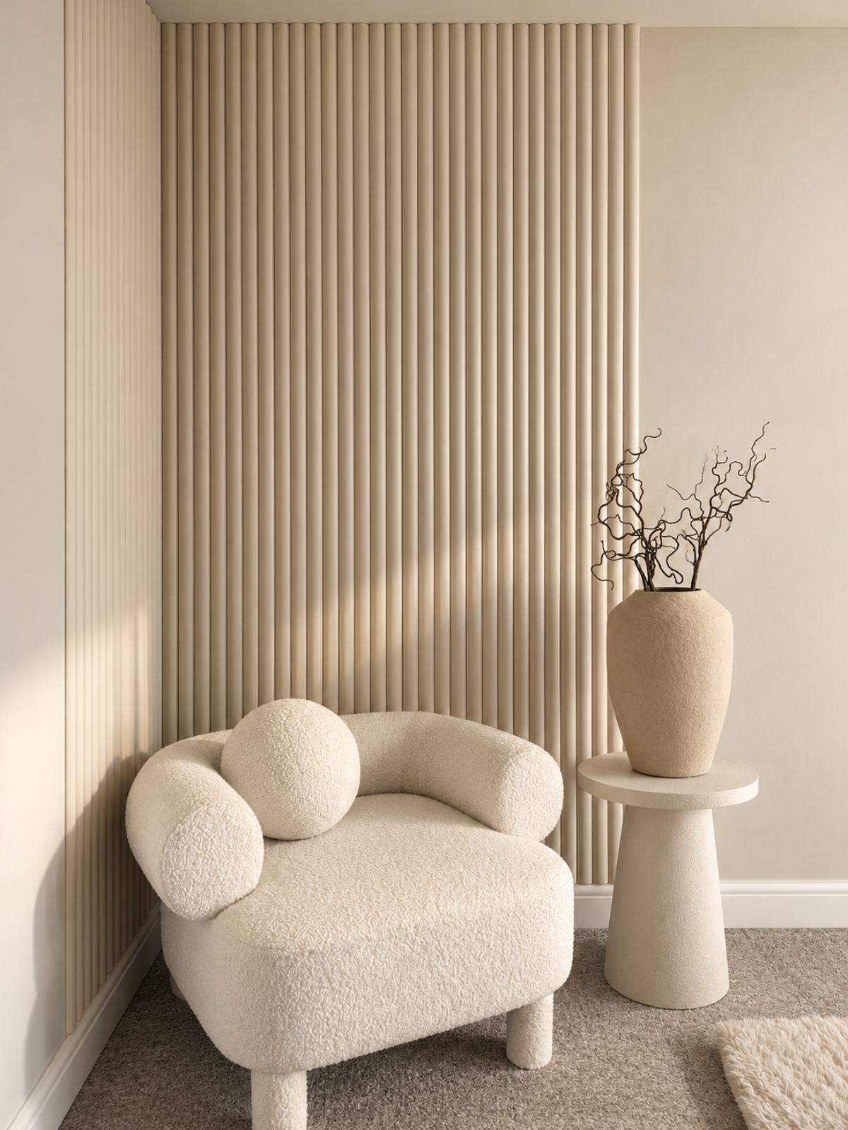 A cream bouclé lounge chair with two round bolsters sits in a corner beside a small round side table holding a tall textured vase with dried branches. The room has warm beige walls, vertical ribbed paneling, and a neutral carpet.