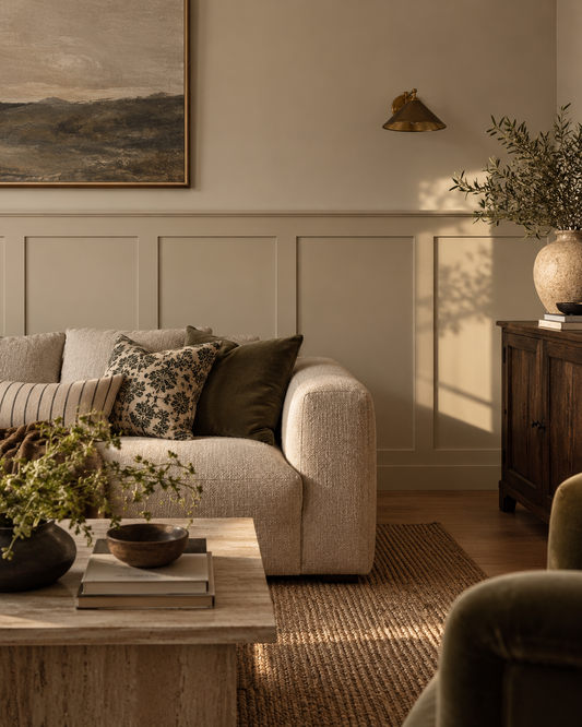 A styled living room scene featuring a cream textured sofa with patterned and olive green pillows, set in a warm, sunlit room with beige shaker wall panelling, a wooden sideboard, and a coffee table in the foreground.