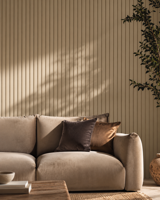 A beige upholstered sofa in a styled living room setting, decorated with two dark brown throw pillows. The background features a beige vertical wood slat wall with soft leaf shadows cast across it, and a potted plant is visible on the right.