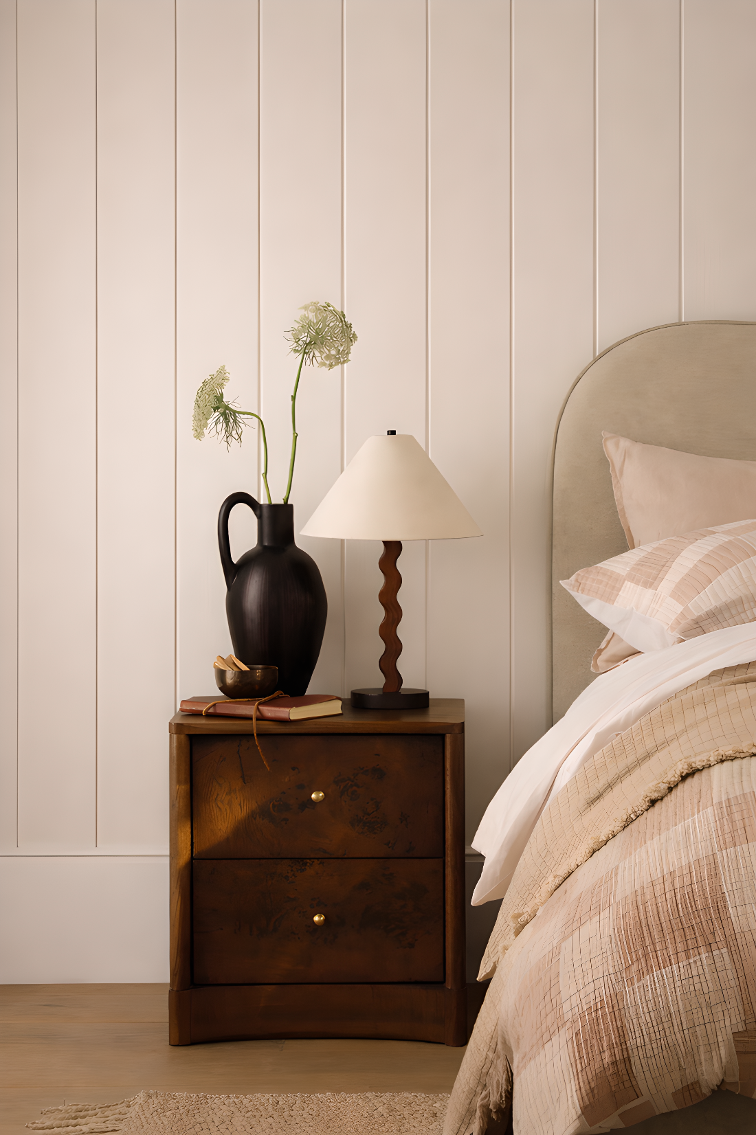 Bedroom with wooden nightstand, lamp, and decorative items against a beige panelled wall.