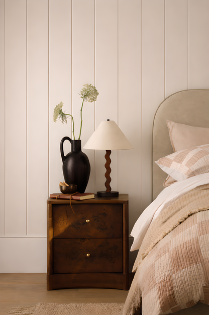 Bedroom with wooden nightstand, lamp, and decorative items against a beige panelled wall.